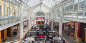 Westgate Arcade in Halifax on a sunny day wit sunlight streaming through the glass panels of the roof and onto tables of diners sitting below